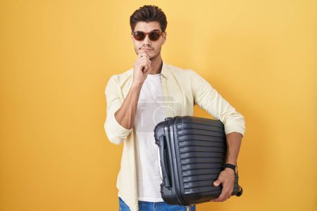 Foto de Young hispanic man holding suitcase going on summer vacation thinking worried about a question, concerned and nervous with hand on chin - Imagen libre de derechos