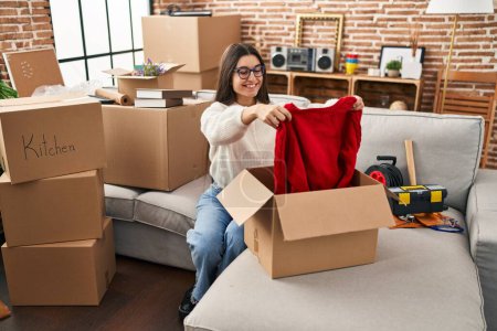 Téléchargez les photos : Young hispanic woman smiling confident unpacking sweater of cardboard box at new home - en image libre de droit