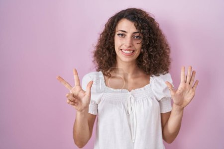 Foto de Mujer hispana con el pelo rizado de pie sobre fondo rosa mostrando y señalando hacia arriba con los dedos número ocho mientras sonríe confiado y feliz. - Imagen libre de derechos