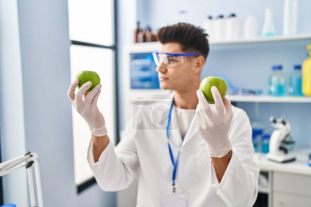 Foto de Young hispanic man wearing scientist uniform holding apples at laboratory - Imagen libre de derechos