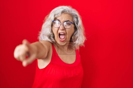 Téléchargez les photos : Femme d'âge moyen avec les cheveux gris debout sur fond rouge pointant mécontent et frustré à la caméra, en colère et furieux contre vous - en image libre de droit