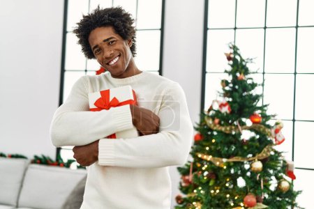 Photo for Young african american man holding gift standing by christmas tree at home. - Royalty Free Image