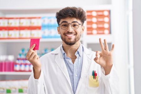 Téléchargez les photos : Arab man with beard working at pharmacy drugstore holding condom smiling looking to the side and staring away thinking. - en image libre de droit