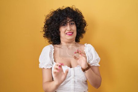 Foto de Young brunette woman with curly hair standing over yellow background disgusted expression, displeased and fearful doing disgust face because aversion reaction. - Imagen libre de derechos