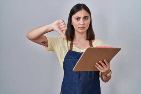 Photo for Hispanic girl wearing apron holding clipboard with angry face, negative sign showing dislike with thumbs down, rejection concept - Royalty Free Image
