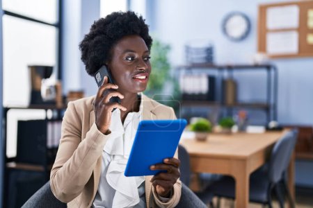 Photo for Young african american woman business worker using touchpad talking on the smartphone at office - Royalty Free Image
