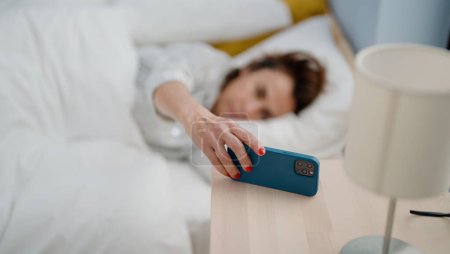 Foto de Mujer hispana de mediana edad apagando la alarma de smartphone acostada en la cama en el dormitorio - Imagen libre de derechos