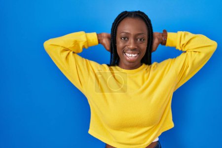 Foto de Beautiful black woman standing over blue background relaxing and stretching, arms and hands behind head and neck smiling happy - Imagen libre de derechos