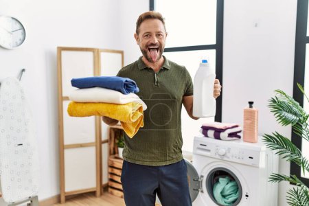 Foto de Middle age man holding clean laundry and detergent bottle sticking tongue out happy with funny expression. - Imagen libre de derechos