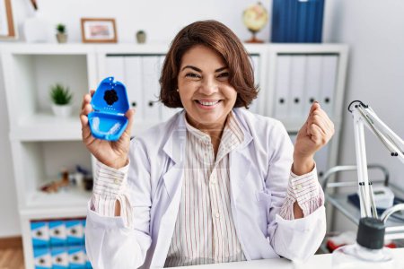 Téléchargez les photos : Dentiste hispanique du Moyen Age femme travaillant à la clinique dentaire tenant aligneur invisible criant fier, célébrant la victoire et le succès très excité avec les bras levés - en image libre de droit