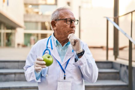 Téléchargez les photos : Médecin sénior avec les cheveux gris tenant pomme verte saine visage sérieux penser à la question avec la main sur le menton, réfléchi à l'idée déroutante - en image libre de droit