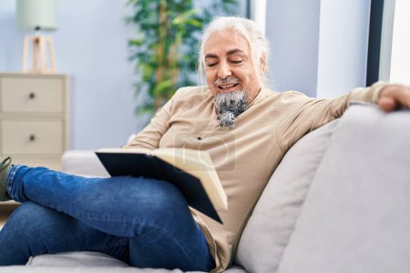 Photo for Middle age grey-haired man reading book sitting on sofa at home - Royalty Free Image