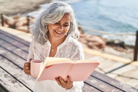 Téléchargez les photos : Femme d'âge moyen lisant le livre et buvant le café assis sur le banc au bord de la mer - en image libre de droit
