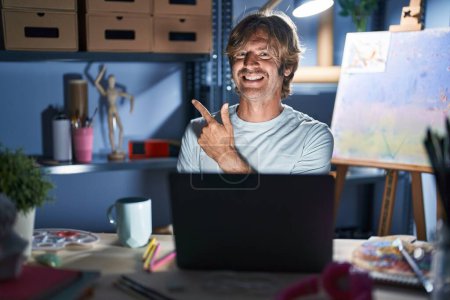 Téléchargez les photos : Middle age man sitting at art studio with laptop at night cheerful with a smile of face pointing with hand and finger up to the side with happy and natural expression on face - en image libre de droit