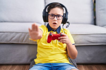 Téléchargez les photos : Jeune enfant hispanique jouant contrôleur de tenue de jeu vidéo portant des écouteurs faisant geste d'arrêt avec les paumes des mains, expression de colère et de frustration - en image libre de droit