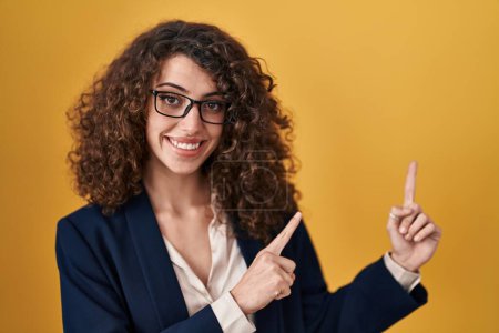 Foto de Hispanic woman with curly hair standing over yellow background smiling and looking at the camera pointing with two hands and fingers to the side. - Imagen libre de derechos
