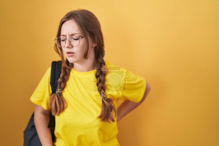Foto de Mujer joven caucásica con mochila de estudiante sobre fondo amarillo sufriendo de dolor de espalda, tocando la espalda con la mano, dolor muscular - Imagen libre de derechos