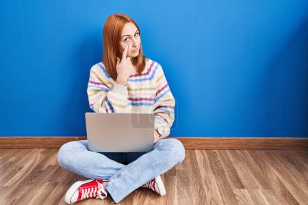 Téléchargez les photos : Young woman using laptop at home sitting on the floor pointing to the eye watching you gesture, suspicious expression - en image libre de droit