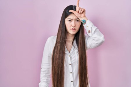 Photo for Chinese young woman standing over pink background making fun of people with fingers on forehead doing loser gesture mocking and insulting. - Royalty Free Image