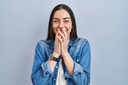 Téléchargez les photos : Femme hispanique debout sur fond bleu rire et rire embarrassé couvrant la bouche avec les mains, commérages et concept scandale - en image libre de droit