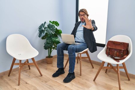 Foto de Hombre guapo de mediana edad sentado en la sala de espera trabajando con el ordenador portátil haciendo gesto de parada con las manos palmas, enojado y expresión de frustración - Imagen libre de derechos