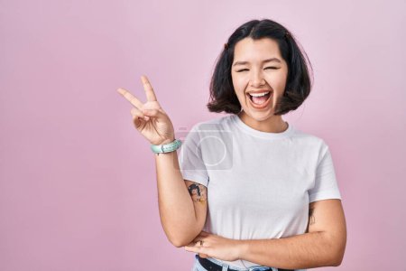 Photo for Young hispanic woman wearing casual white t shirt over pink background smiling with happy face winking at the camera doing victory sign with fingers. number two. - Royalty Free Image
