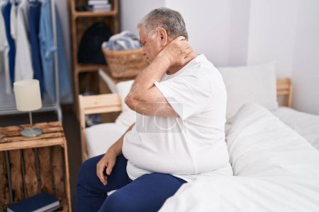 Téléchargez les photos : Homme d'âge moyen aux cheveux gris souffrant d'une blessure au cou assis sur le lit dans la chambre - en image libre de droit