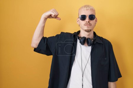 Téléchargez les photos : Jeune homme caucasien portant des lunettes de soleil debout sur fond jaune personne forte montrant muscle du bras, confiant et fier de la puissance - en image libre de droit