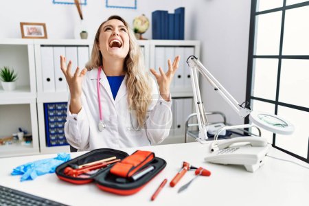 Photo for Young beautiful doctor woman with reflex hammer and medical instruments crazy and mad shouting and yelling with aggressive expression and arms raised. frustration concept. - Royalty Free Image