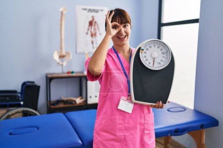 Photo for Young brunette woman as nutritionist holding weighing machine smiling happy doing ok sign with hand on eye looking through fingers - Royalty Free Image