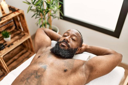 Téléchargez les photos : Jeune homme afro-américain détendu avec les mains sur la tête allongée sur une table de massage au centre de beauté - en image libre de droit