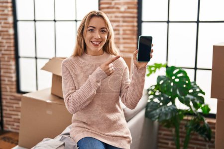 Photo for Hispanic woman moving to a new home showing smartphone screen smiling happy pointing with hand and finger - Royalty Free Image
