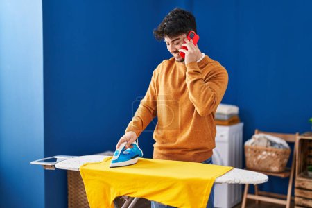 Téléchargez les photos : Young hispanic man talking on the smartphone ironing clothes at laundry room - en image libre de droit
