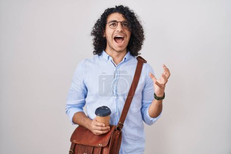 Foto de Hombre hispano con el pelo rizado bebiendo una taza de café para llevar loco y loco gritando y gritando con expresión agresiva y los brazos levantados. concepto de frustración. - Imagen libre de derechos