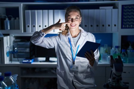 Photo for Beautiful blonde woman working at scientist laboratory late at night pointing with hand finger to face and nose, smiling cheerful. beauty concept - Royalty Free Image