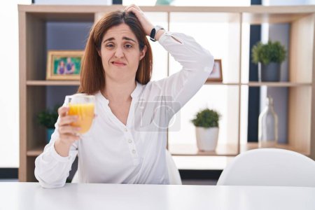 Foto de Brunette woman drinking glass of orange juice confuse and wondering about question. uncertain with doubt, thinking with hand on head. pensive concept. - Imagen libre de derechos
