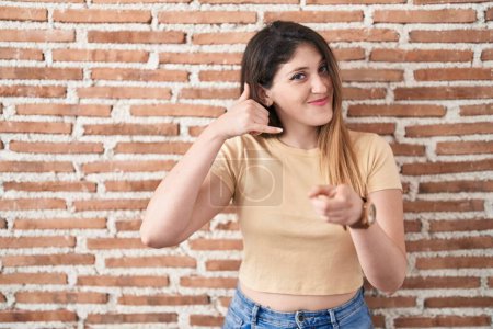 Téléchargez les photos : Jeune femme brune debout au-dessus du mur de briques souriant faisant parler au téléphone geste et vous pointant du doigt. Appelle-moi. - en image libre de droit