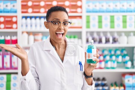Téléchargez les photos : Femme afro-américaine travaillant à la pharmacie drugstore tenant sirop célébrant la réalisation avec sourire heureux et expression gagnante avec la main levée - en image libre de droit