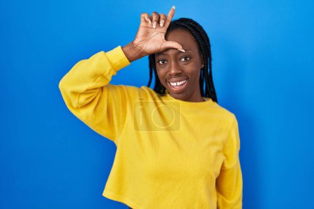 Photo for Beautiful black woman standing over blue background making fun of people with fingers on forehead doing loser gesture mocking and insulting. - Royalty Free Image