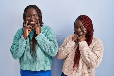 Téléchargez les photos : Two african woman standing over blue background smiling with open mouth, fingers pointing and forcing cheerful smile - en image libre de droit