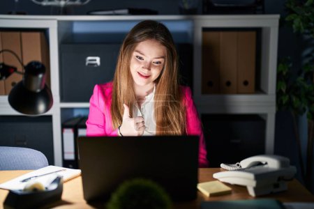 Foto de Young caucasian woman working at the office at night doing happy thumbs up gesture with hand. approving expression looking at the camera showing success. - Imagen libre de derechos