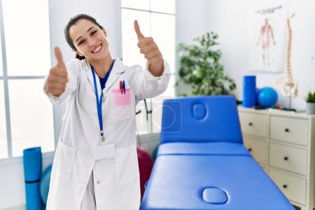 Photo for Young doctor woman working at pain recovery clinic approving doing positive gesture with hand, thumbs up smiling and happy for success. winner gesture. - Royalty Free Image