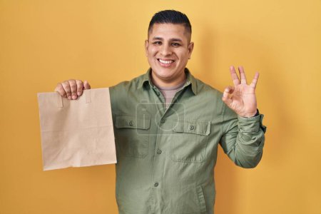 Foto de Joven hispano sosteniendo llevar bolsa de papel haciendo signo ok con los dedos, sonriendo gesto amistoso excelente símbolo - Imagen libre de derechos
