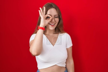 Téléchargez les photos : Jeune femme caucasienne debout sur fond rouge faisant un geste correct avec la main souriante, les yeux regardant à travers les doigts avec un visage heureux. - en image libre de droit