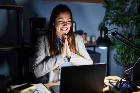 Téléchargez les photos : Jeune femme brune travaillant au bureau la nuit priant avec les mains ensemble demandant pardon en souriant confiant. - en image libre de droit