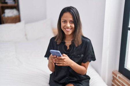Téléchargez les photos : Young hispanic woman using smartphone sitting on bed at bedroom - en image libre de droit