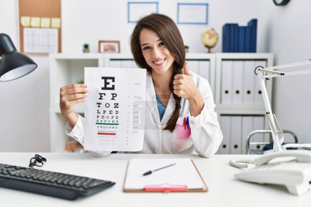 Photo for Young doctor woman holding eyesight test smiling happy and positive, thumb up doing excellent and approval sign - Royalty Free Image