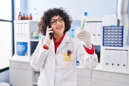 Téléchargez les photos : Jeune belle femme hispanique scientifique parlant sur smartphone tenant le tube à essai au laboratoire - en image libre de droit