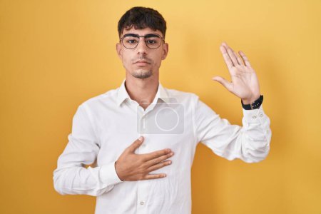 Foto de Joven hombre hispano de pie sobre fondo amarillo jurando con la mano en el pecho y la palma abierta, haciendo un juramento de lealtad promesa - Imagen libre de derechos