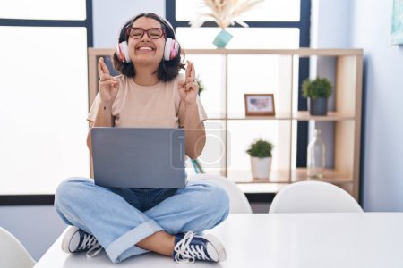 Foto de Young hispanic woman using laptop sitting on the table wearing headphones gesturing finger crossed smiling with hope and eyes closed. luck and superstitious concept. - Imagen libre de derechos